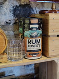 A Rum Lover's Puzzle placed on a wooden shelf next to a clear glass, with a rustic stone wall, wooden crates, and nautical decor in the background.