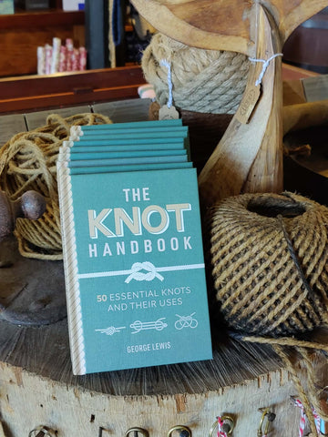 The Knot Handbook book by George Lewis standing upright on a rustic wooden stump, surrounded by coiled twine, rope, and wooden nautical decor in a styled indoor setting.